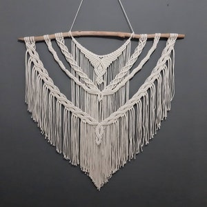 May include: A white macrame wall hanging with a wooden dowel at the top. The hanging features a geometric pattern with fringe.