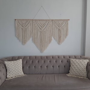 Pode incluir: Uma tapeçaria de macramê bege com três seções triangulares, cada uma com um padrão diferente. A tapeçaria está presa a uma haste de madeira e está pendurada em uma parede branca acima de um sofá cinza com duas almofadas de macramê bege.