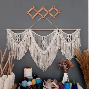 Pode incluir: Uma tapeçaria de macramé branca com uma barra de madeira, que apresenta um padrão geométrico com franjas. A tapeçaria de macramé é exibida em uma parede cinza com um cabide de madeira acima dela. Há vários rolos de fio coloridos e uma caixa de madeira em uma mesa branca na frente da tapeçaria de macramé.