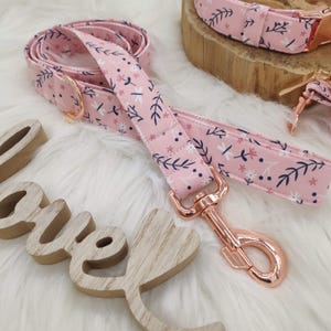 Könnte beinhalten: Eine rosa Hunde-Leine mit einem Blumenmuster und einem roségoldenen Metallverschluss. Die Leine ist aus Stoff und hat einen weißen Hintergrund mit rosa Blumen und dunkelblauen Blättern.