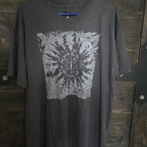 Può includere: Una t-shirt a maniche corte grigio antracite con una stampa grafica grigio chiaro raffigurante un occhio centrale circondato da pini, funghi e il testo "Be the Cryptid You Want to See in the Woods." La maglietta ha un girocollo.