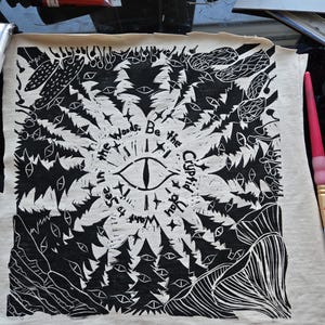 May include: Black and white linocut print on fabric featuring a central eye design surrounded by trees and text that reads "The Woods. Be the Cupid You Want to See In". The print has a rustic, handmade aesthetic.