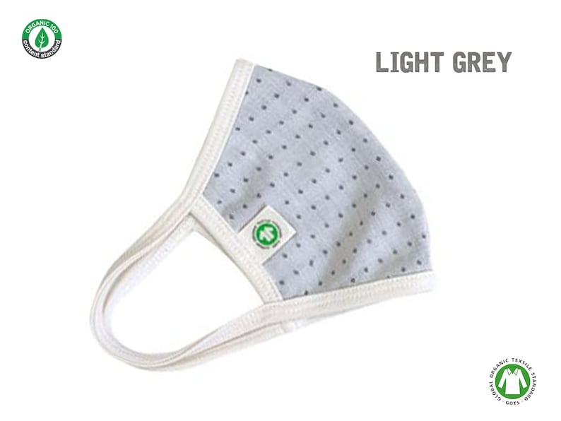 May include: Light grey face mask with white trim and small white polka dots. The mask has a white elastic band for the ears. The mask has a green logo with the text "Organic 100" and a leaf.