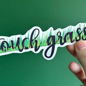 Puede incluir: Una pegatina blanca con la frase "touch grass" en escritura cursiva negra. Las letras están sobre un fondo de hierba verde y amarilla. La pegatina se sostiene sobre un fondo verde.