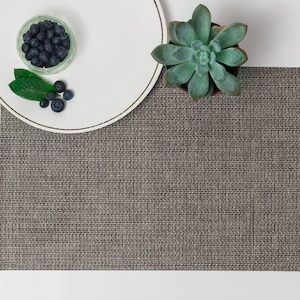 Puede incluir: Un mantel individual tejido gris con un plato blanco y un pequeño cuenco de vidrio con arándanos en la parte superior. Una pequeña planta suculenta verde se encuentra a la derecha del plato.