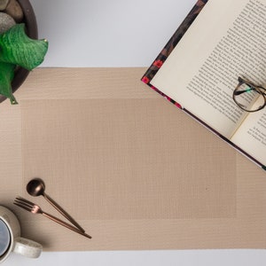 Puede incluir: Un mantel individual beige con textura tejida, una taza blanca, un tenedor, una cuchara y un par de gafas sobre un libro abierto.