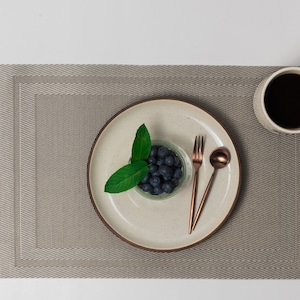 Puede incluir: Un plato de cerámica blanco con un pequeño cuenco de vidrio de arándanos y una ramita de menta verde. Un tenedor y una cuchara de oro rosa están en el plato. Una taza blanca con café negro está en un mantel individual marrón.