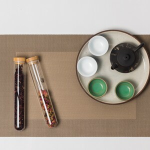 Puede incluir: Un juego de té con una tetera negra, tazas blancas y verdes, y tubos de vidrio llenos de hojas de té y pétalos de rosa. El juego está sobre un mantel individual marrón, creando un ambiente sereno.
