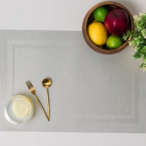 Puede incluir: Un mantel individual gris claro con una superficie texturizada, un tenedor y una cuchara dorados, y un vaso de agua con una rodaja de limón. Un cuenco de madera contiene una manzana roja, un limón y dos limas. También hay una pequeña planta verde.