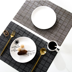 Puede incluir: Dos manteles individuales, uno gris y otro gris oscuro, cada uno con un patrón de cuadrícula. Un plato blanco está sobre el mantel individual gris, y un segundo plato blanco con texto y un sol está sobre el mantel individual gris oscuro. Tenedor, cuchara y taza de café dorados.