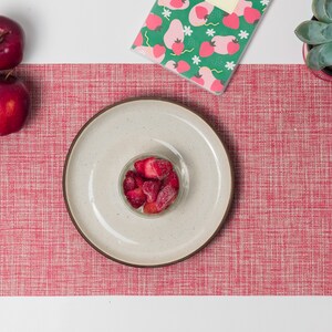 Puede incluir: Vista aérea de una mesa con un mantel individual tejido rojo. Un plato beige con borde marrón contiene un pequeño cuenco de cristal lleno de fresas en rodajas. También hay dos manzanas rojas y un cuaderno verde con un estampado de fresas.