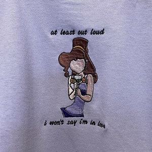 Może przedstawiać: Koszulka w kolorze lawendowym z haftowanym obrazem kobiety trzymającej kwiat. Tekst "at least out loud" i "I won't say I'm in love" jest wyszyty pod obrazem.