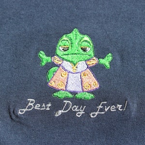 Op de afbeelding: Een donkerblauw T-shirt met een geborduurde groene kameleon die een paars-gouden jurk draagt. De tekst "Best Day Ever!" is onder de kameleon geborduurd.