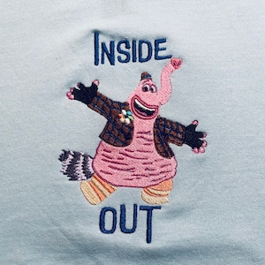 May include: Light blue embroidered sweatshirt featuring the words "INSIDE OUT" in dark blue. The design includes a pink elephant-like character with a brown jacket, black gloves, and colorful socks. The character has its arms open.