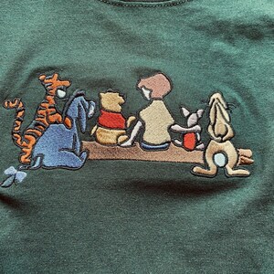 May include: Green t-shirt with an embroidered image of Winnie the Pooh and his friends, including Tigger, Piglet, Eeyore, and Rabbit. The characters are sitting on a log and smiling.