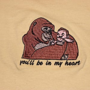 Puede incluir: Dise&ntilde;o bordado en una camiseta amarilla con un gorila marr&oacute;n sosteniendo un peque&ntilde;o mono beb&eacute; rosa. El texto "you'll be in my heart" est&aacute; bordado debajo de la imagen.