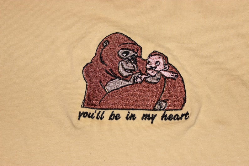Puede incluir: Dise&ntilde;o bordado en una camiseta amarilla con un gorila marr&oacute;n sosteniendo un peque&ntilde;o mono beb&eacute; rosa. El texto "you'll be in my heart" est&aacute; bordado debajo de la imagen.