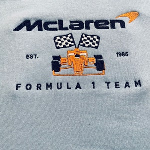 Op de afbeelding: Lichtblauw sweatshirt met geborduurd "McLaren" logo, een Formule 1 raceauto en geblokte vlaggen. De tekst "EST. 1985" en "FORMULA 1 TEAM" zijn ook geborduurd.