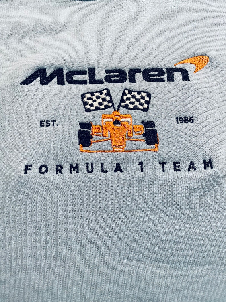 Op de afbeelding: Lichtblauw sweatshirt met geborduurd "McLaren" logo, een Formule 1 raceauto en geblokte vlaggen. De tekst "EST. 1985" en "FORMULA 1 TEAM" zijn ook geborduurd.