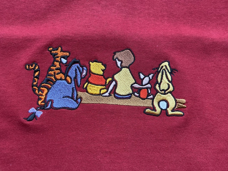May include: Embroidered image of Winnie the Pooh and his friends Tigger, Piglet, Eeyore, and Rabbit. The characters are all sitting together on a brown log. The image is on a red background.