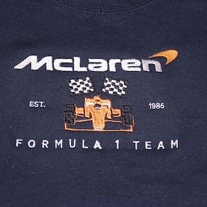 Op de afbeelding: Marineblauw sweatshirt met geborduurd "McLaren" logo, oranje en zwarte raceauto afbeelding en "FORMULA 1 TEAM" tekst. Voorzien van de tekst "EST. 1985".
