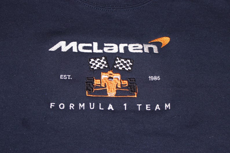 Op de afbeelding: Marineblauw sweatshirt met geborduurd "McLaren" logo, oranje en zwarte raceauto afbeelding en "FORMULA 1 TEAM" tekst. Voorzien van de tekst "EST. 1985".