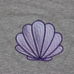 May include: A gray fabric with a detailed embroidered seashell design. The seashell is in shades of purple with a dark purple outline. The design is centered on the fabric, showcasing a coastal or nautical theme.