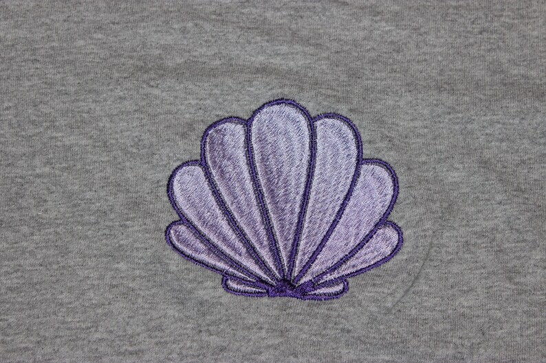 May include: A gray fabric with a detailed embroidered seashell design. The seashell is in shades of purple with a dark purple outline. The design is centered on the fabric, showcasing a coastal or nautical theme.