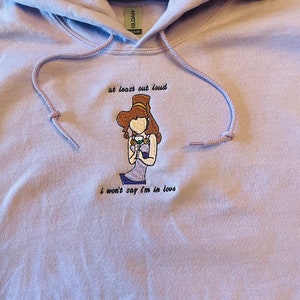 May include: A lavender hoodie with an embroidered image of a woman wearing a crown and holding a glass of wine. The text "at least out loud" is above the image and the text "i won't say i'm in love" is below the image.