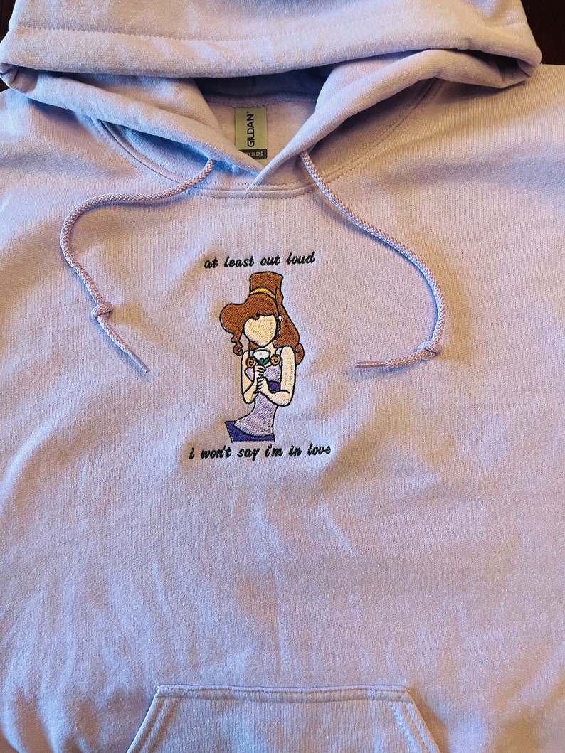 May include: A lavender hoodie with an embroidered image of a woman wearing a crown and holding a glass of wine. The text "at least out loud" is above the image and the text "i won't say i'm in love" is below the image.