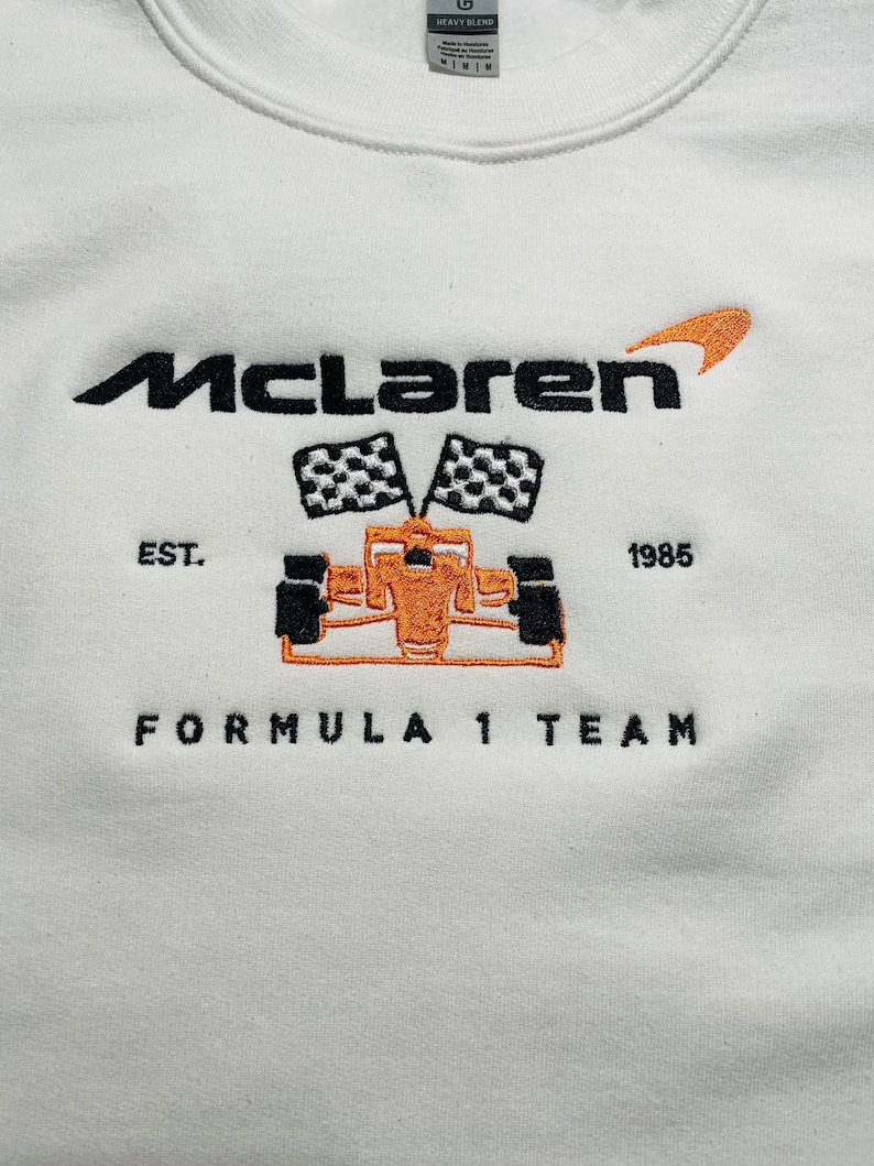 Op de afbeelding: Wit sweatshirt met geborduurd McLaren Formula 1 Team-ontwerp. Voorzien van het McLaren-logo, een raceauto, geblokte vlaggen en de tekst "EST. 1985". Het ontwerp is zwart en oranje.