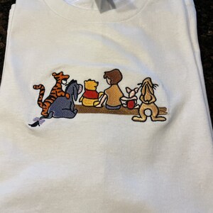 May include: White t-shirt with an embroidered image of Winnie the Pooh, Piglet, Tigger, Eeyore, and Christopher Robin. The characters are sitting on a log.