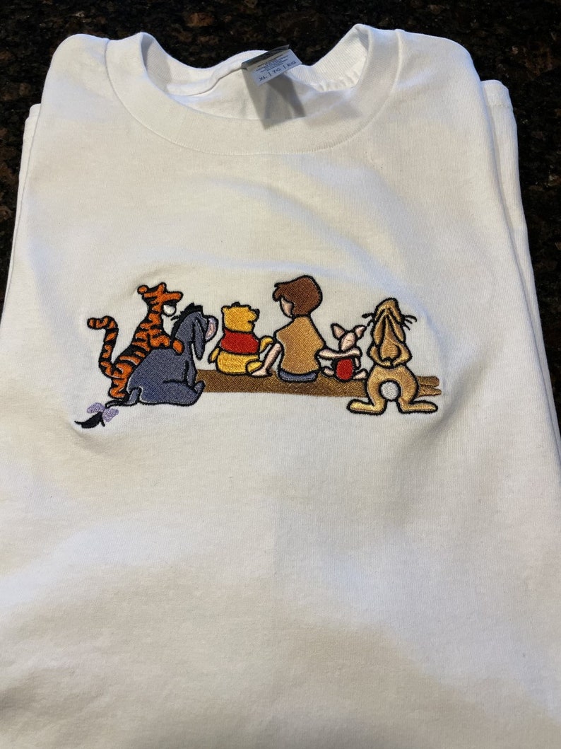 May include: White t-shirt with an embroidered image of Winnie the Pooh, Piglet, Tigger, Eeyore, and Christopher Robin. The characters are sitting on a log.