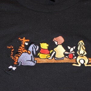May include: Embroidered black t-shirt featuring Winnie the Pooh and his friends Piglet, Tigger, and Eeyore sitting on a log.