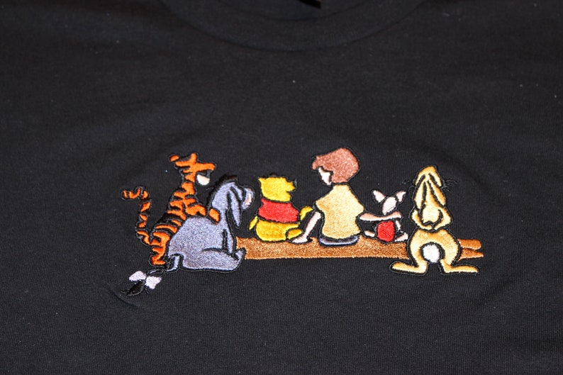 May include: Embroidered black t-shirt featuring Winnie the Pooh and his friends Piglet, Tigger, and Eeyore sitting on a log.