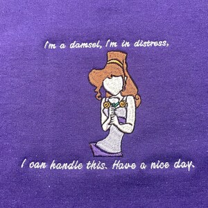 May include: Purple sweatshirt with white embroidered text that reads "I'm a damsel, I'm in distress, I can handle this. Have a nice day." and an embroidered image of a cartoon woman holding a flower.