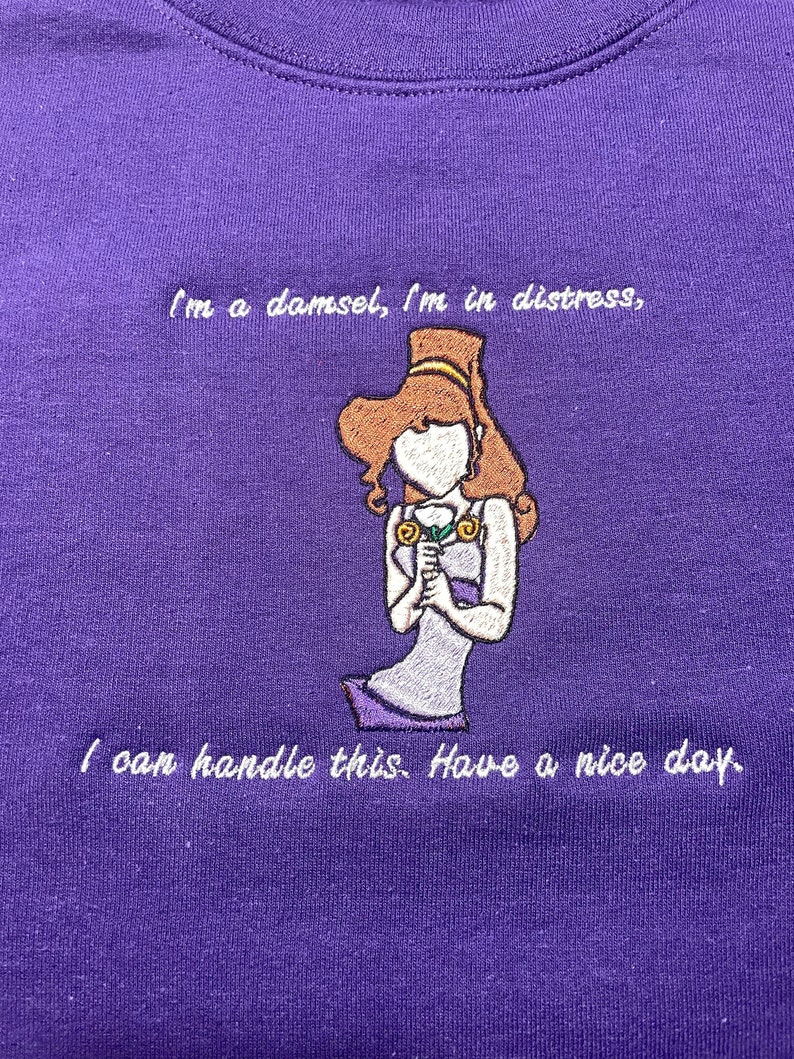 May include: Purple sweatshirt with white embroidered text that reads "I'm a damsel, I'm in distress, I can handle this. Have a nice day." and an embroidered image of a cartoon woman holding a flower.