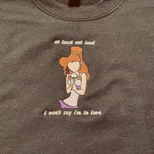 May include: A black sweatshirt with an embroidered image of a woman with brown hair and a purple dress. The text "at least out loud I won't say I'm in love" is embroidered below the image.