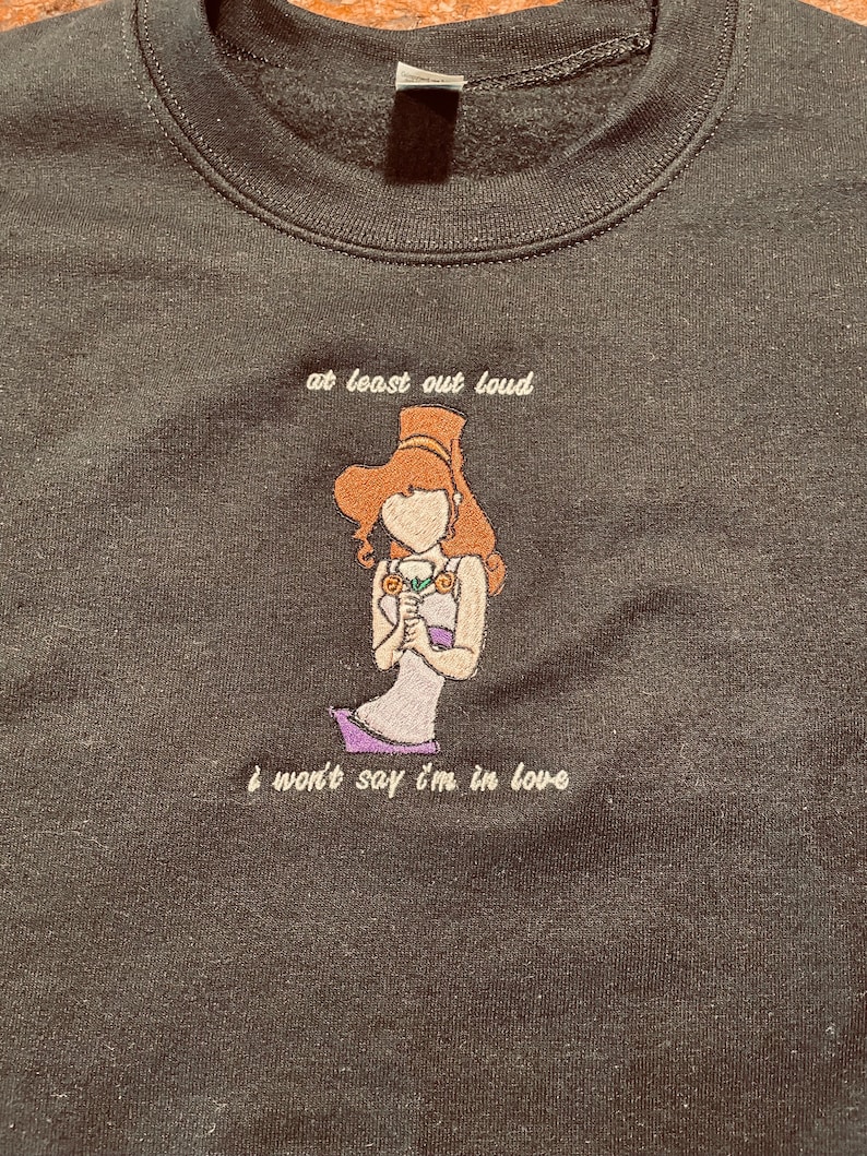 May include: A black sweatshirt with an embroidered image of a woman with brown hair and a purple dress. The text "at least out loud I won't say I'm in love" is embroidered below the image.