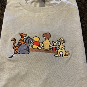 May include: A light gray t-shirt with an embroidered image of Winnie the Pooh and his friends. The characters are Tigger, Piglet, Eeyore, Pooh Bear, and Christopher Robin. The characters are all sitting on a log.