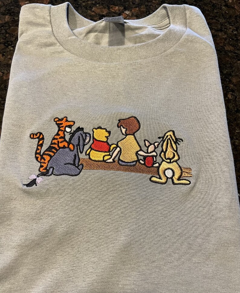 May include: A light gray t-shirt with an embroidered image of Winnie the Pooh and his friends. The characters are Tigger, Piglet, Eeyore, Pooh Bear, and Christopher Robin. The characters are all sitting on a log.