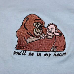 Puede incluir: Dise&ntilde;o bordado en una camiseta azul claro con un gorila marr&oacute;n sosteniendo una peque&ntilde;a figura humana. El texto "you'll be in my heart" est&aacute; debajo de la imagen.