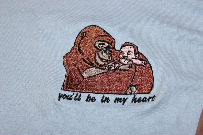 Puede incluir: Dise&ntilde;o bordado en una camiseta azul claro con un gorila marr&oacute;n sosteniendo una peque&ntilde;a figura humana. El texto "you'll be in my heart" est&aacute; debajo de la imagen.