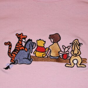 May include: Embroidered design on a pink t-shirt featuring Winnie the Pooh and his friends: Tigger, Piglet, Eeyore, and Rabbit. The characters are sitting on a branch, smiling and looking to the right.