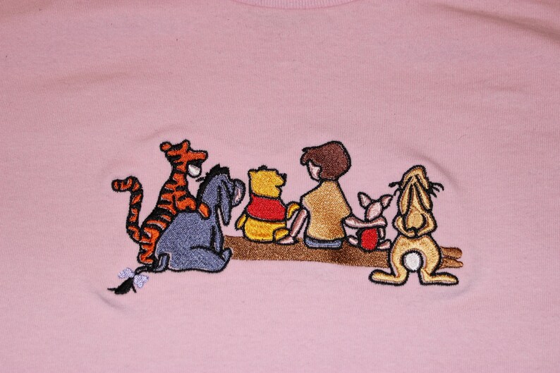 May include: Embroidered design on a pink t-shirt featuring Winnie the Pooh and his friends: Tigger, Piglet, Eeyore, and Rabbit. The characters are sitting on a branch, smiling and looking to the right.