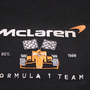 Op de afbeelding: Zwarte sweatshirt met het McLaren-logo in wit en oranje, en een geborduurd Formule 1-raceauto-ontwerp. De tekst "EST. 1986" en "FORMULA 1 TEAM" zijn ook op het kledingstuk geborduurd.