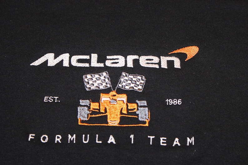 Op de afbeelding: Zwarte sweatshirt met het McLaren-logo in wit en oranje, en een geborduurd Formule 1-raceauto-ontwerp. De tekst "EST. 1986" en "FORMULA 1 TEAM" zijn ook op het kledingstuk geborduurd.