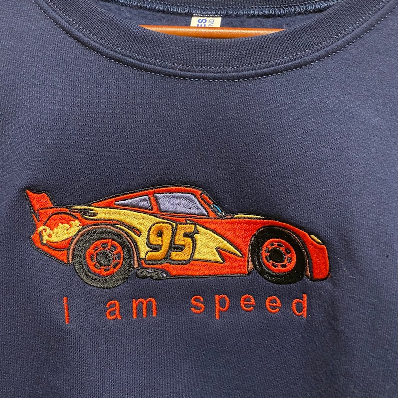 May include: A navy blue sweatshirt with an embroidered image of Lightning McQueen from the Disney Pixar movie Cars. The car is red, yellow, and black with the number 95 on the side. The text "I am speed" is embroidered below the car.