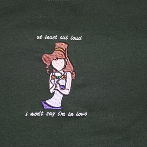 May include: A dark green t-shirt with an embroidered image of a woman holding a flower. The text "at least out loud" and "i won't say i'm in love" is embroidered on the shirt.