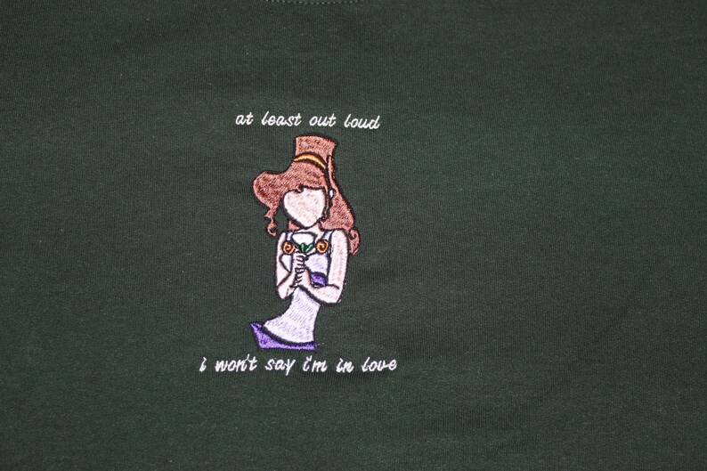 May include: A dark green t-shirt with an embroidered image of a woman holding a flower. The text "at least out loud" and "i won't say i'm in love" is embroidered on the shirt.